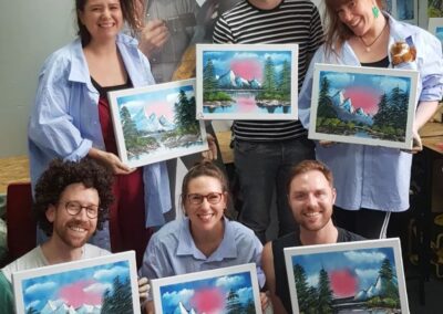 6 people smiling holding their own paintings next to a life size cut out of Bob Ross